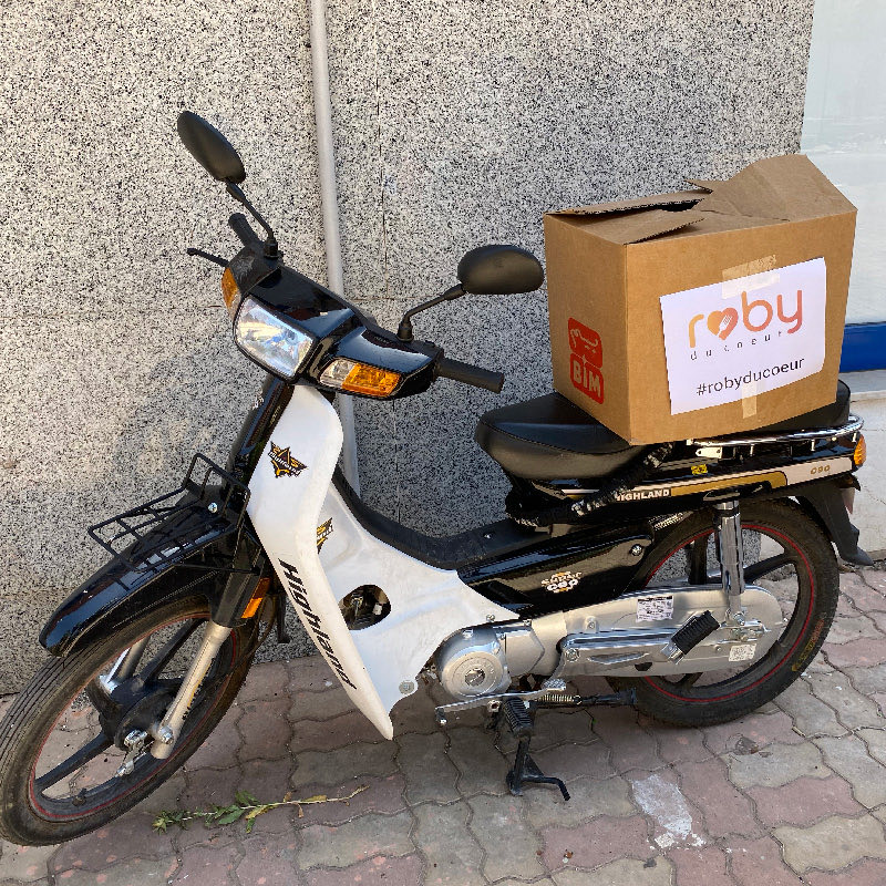 A moped with a Roby du Coeur box strapped to the luggage rack, ready to set off on a delivery.
